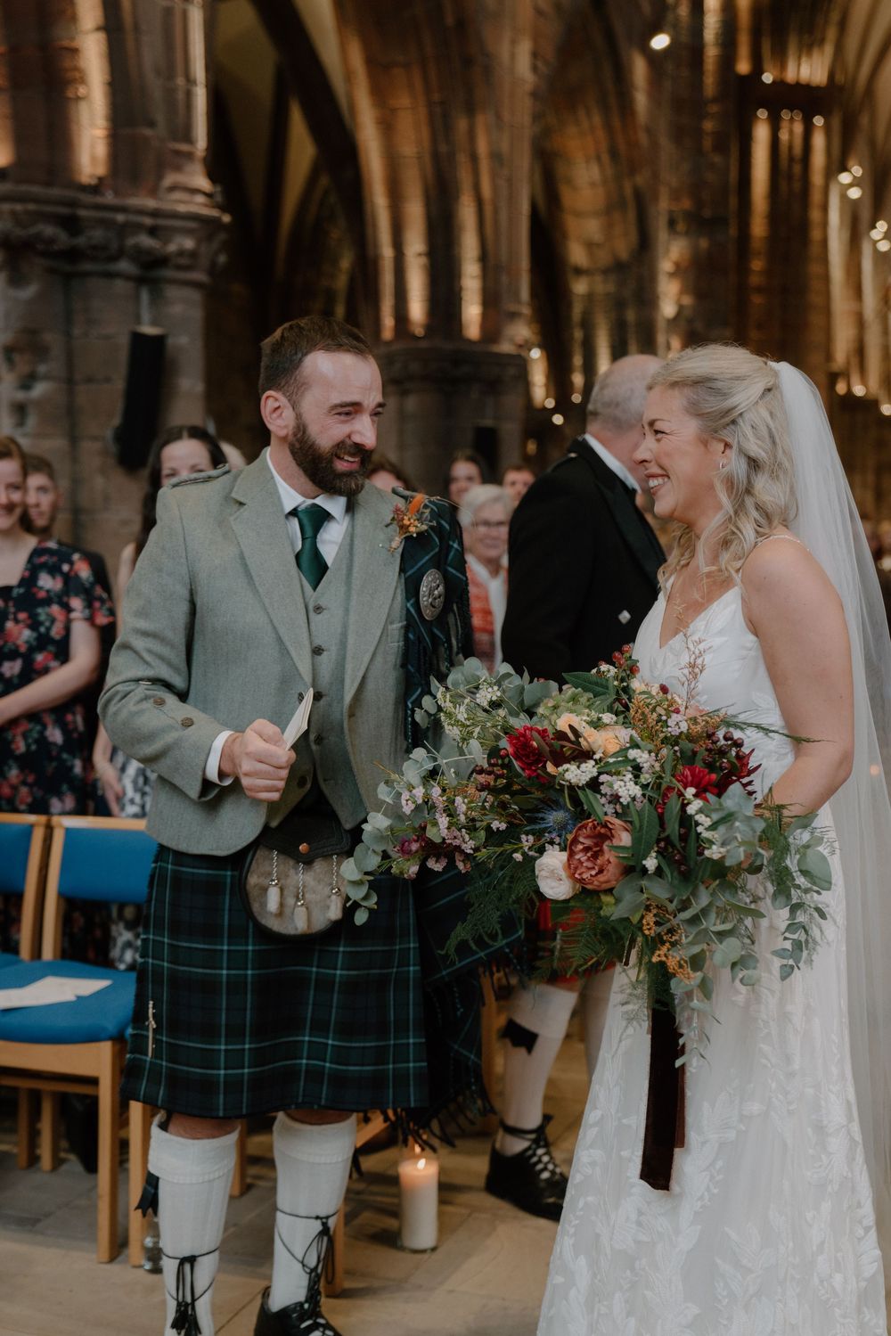 Smiling bride holding lush floral bouquet walks with groom in Scottish tartan kilt inside a stone church.