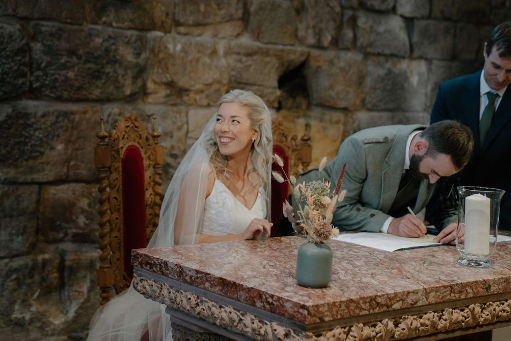 Smiling bride in white dress and veil signs wedding register at ornate table in stone venue.
