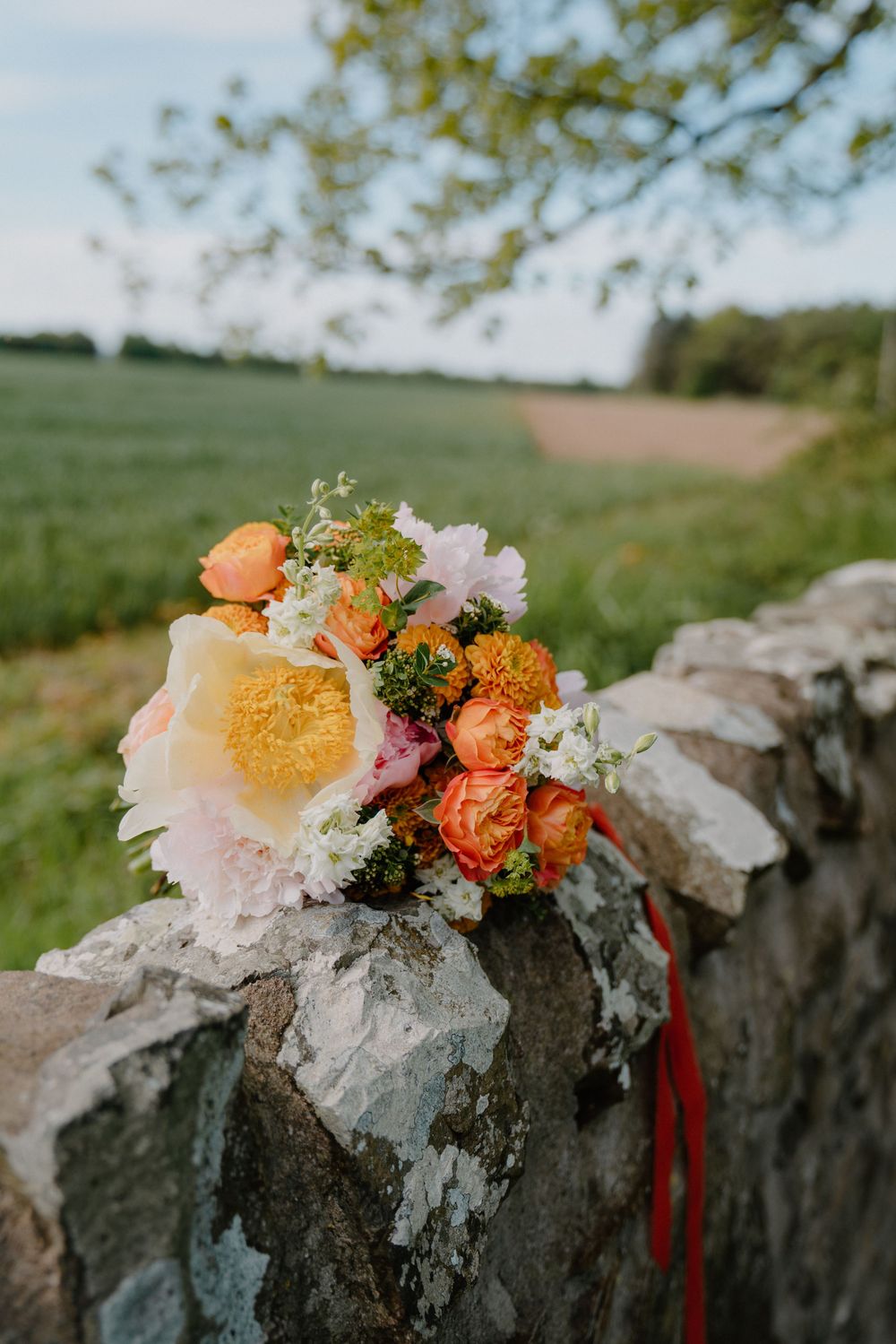 Colorful wedding bouquet with orange, pink, and yellow flowers resting on a rustic stone wall in a green countryside.