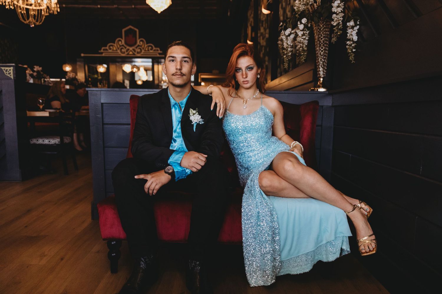 Elegant couple in formal attire posed on vintage furniture in dark venue setting.