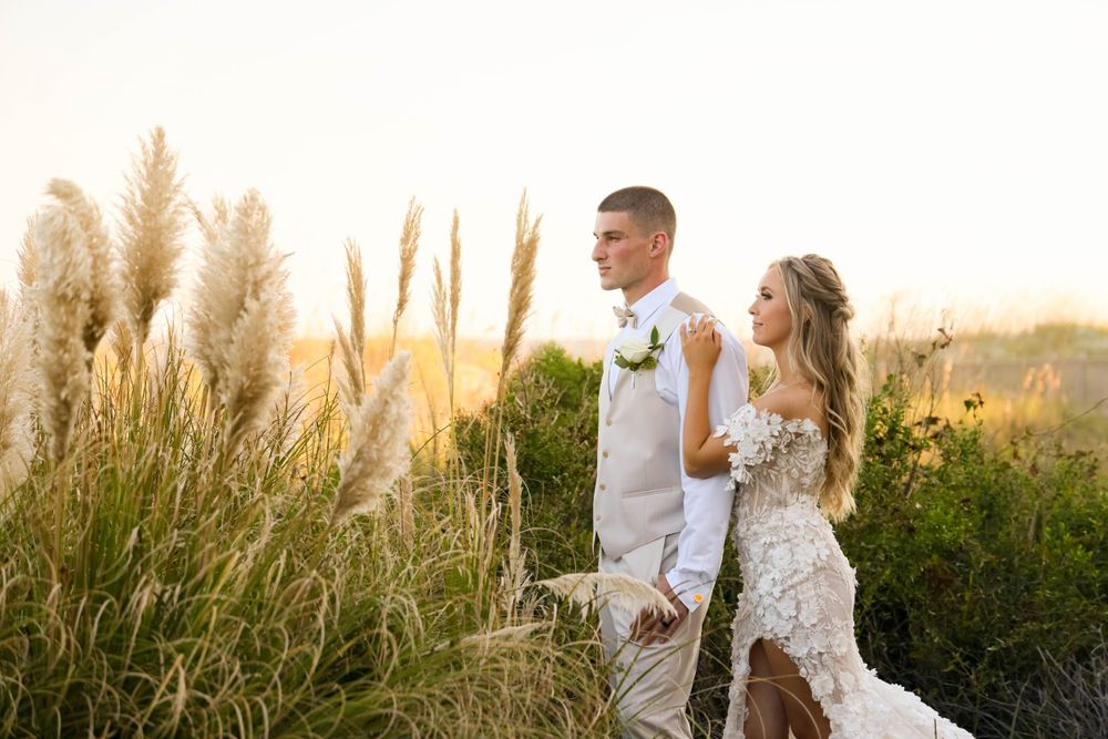 Couple stands among pampas grass at sunset in wedding attire.