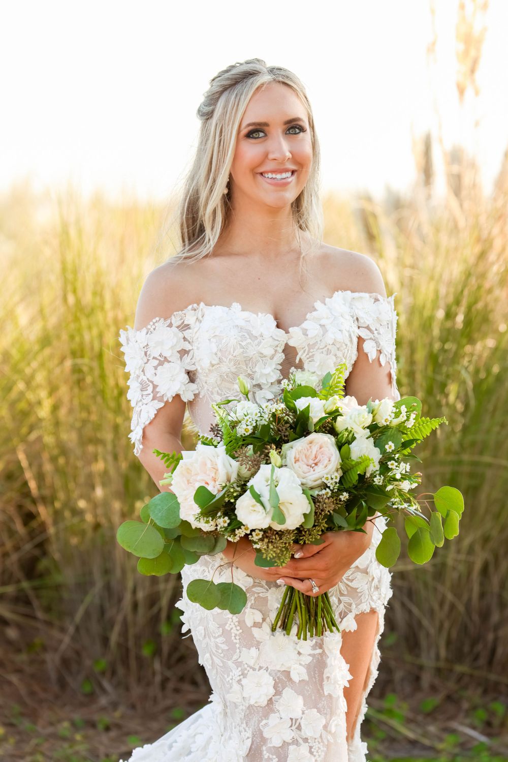 Beautiful bridal portrait in a golden field at sunset featuring an off-shoulder lace dress and white floral bouquet.