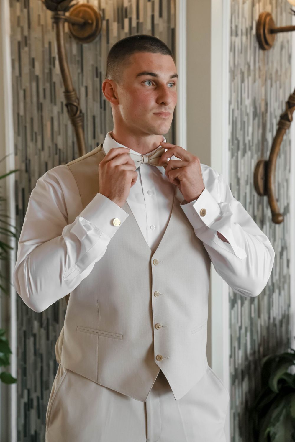 A groom adjusts his bowtie while getting ready in a white suit and vest.