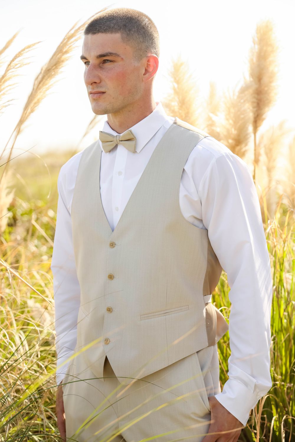 Groom in light colored vest and bow tie poses in tall grass at golden hour.