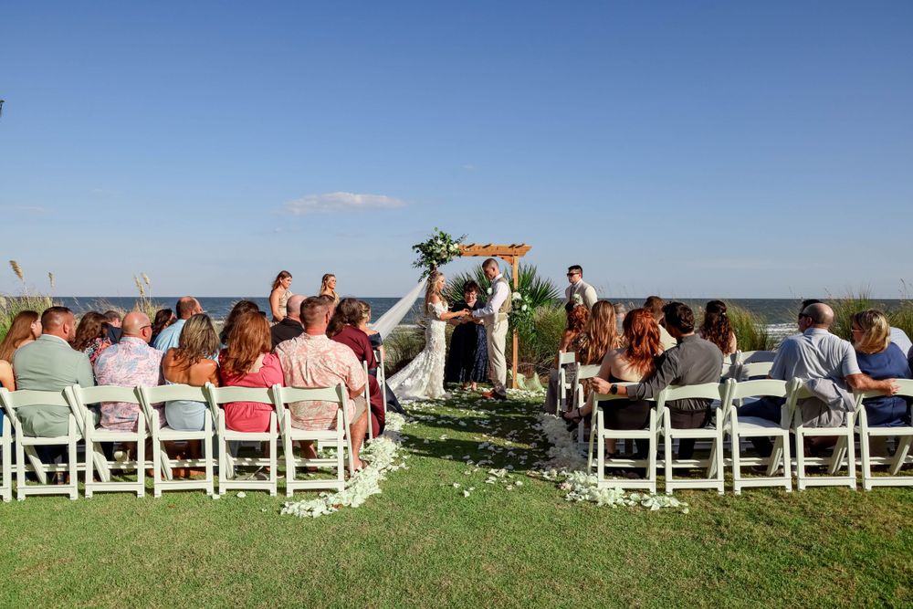 Wedding ceremony in progress at outdoor venue with guests seated on white chairs.