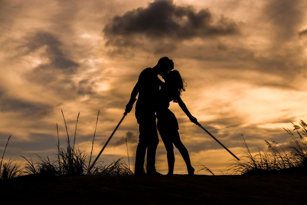 Silhouettes of a couple embracing against a dramatic sunset sky holding lightsabers