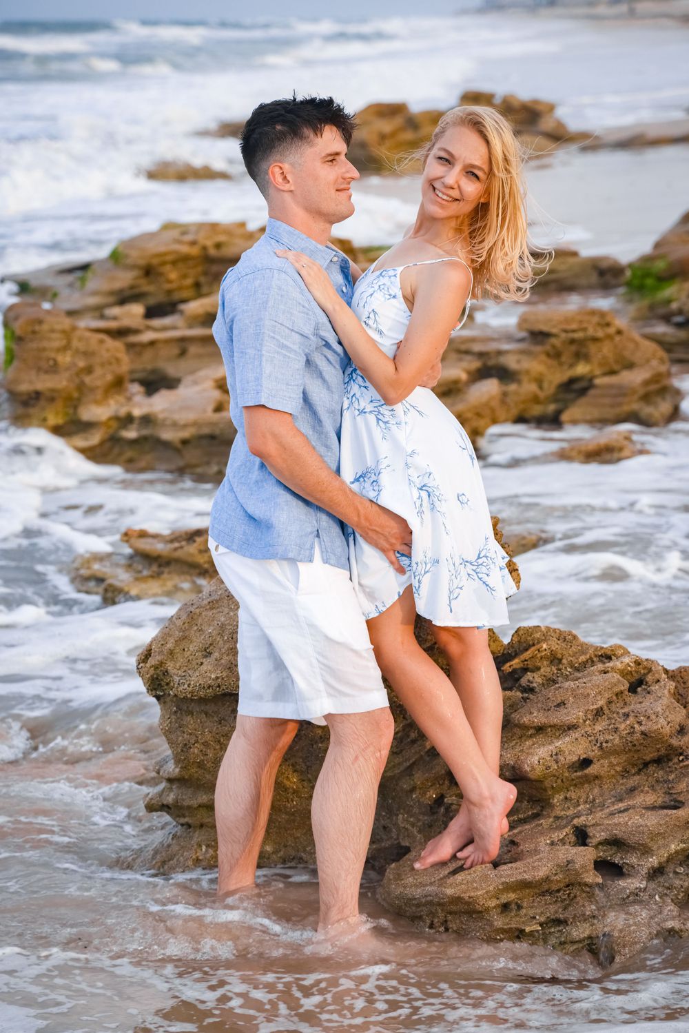 Romantic couple posing on rocky beach in matching blue and white outfits.