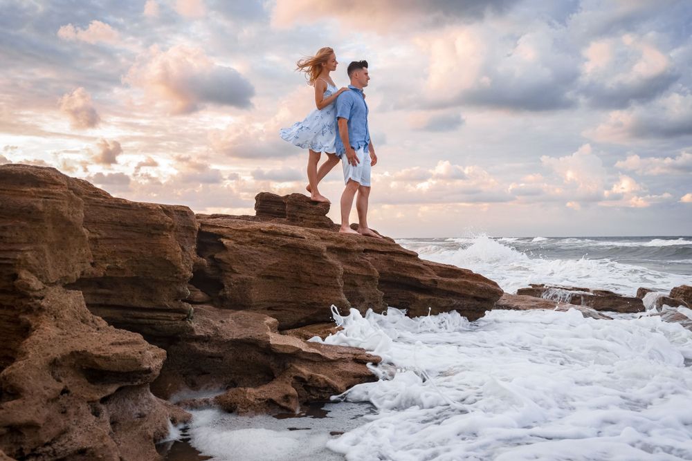 A couple stands on coastal rocks as waves crash below during sunset.