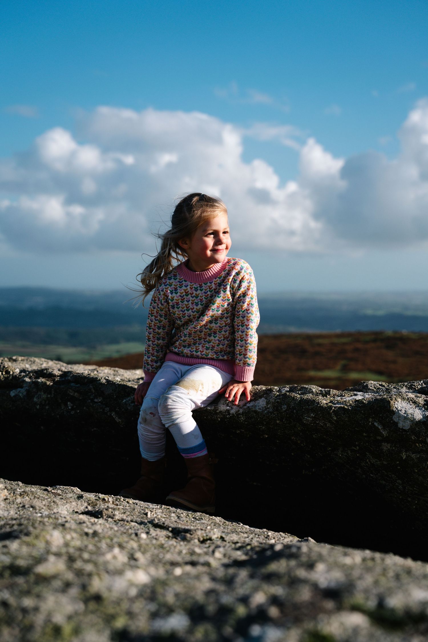 A child in a patterned sweater sits on a rocky ledge overlooking a vast countryside vista.