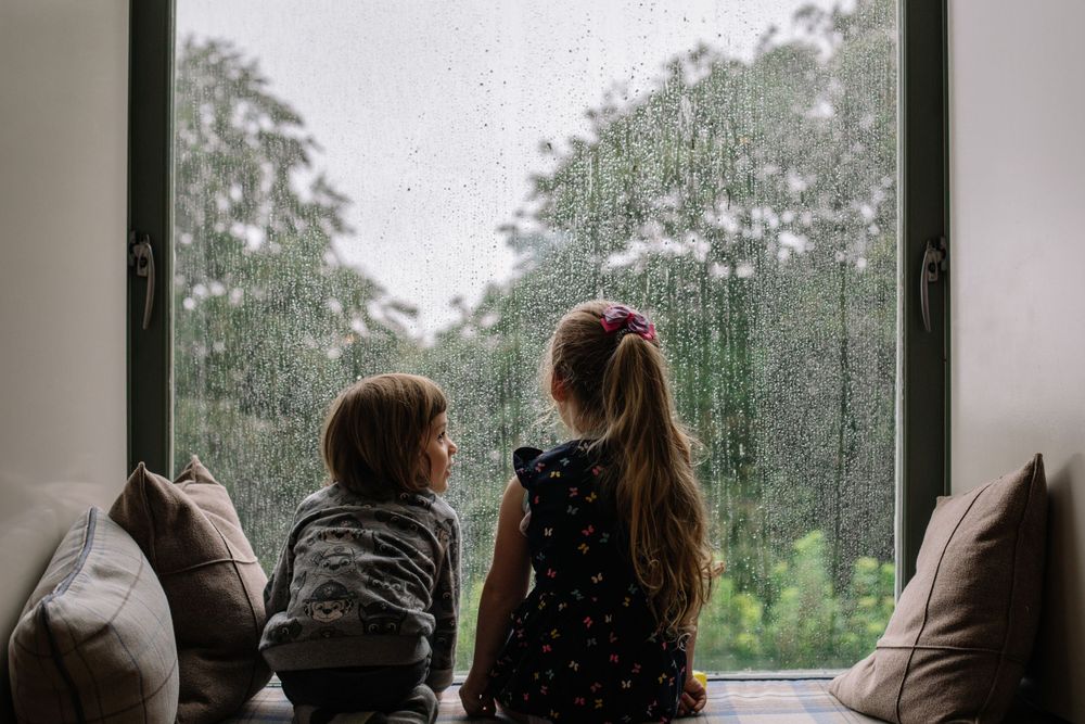 Two children watching rain fall on window while sitting on windowsill cushions.