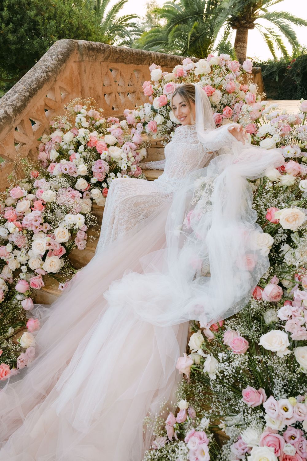 Bride in a flowing lace wedding gown and veil poses on flower-lined stone steps surrounded by pink and white roses.
