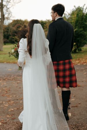 Back view of bride in flowing veil and groom in red tartan kilt walking autumn path.