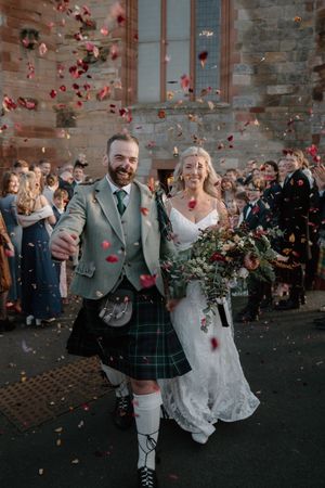 Smiling newlyweds exit church in confetti shower, groom in green tartan kilt, bride in lace gown holding bouquet.