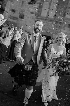 Black and white photo of a smiling couple in wedding attire walking through confetti outside a stone church.