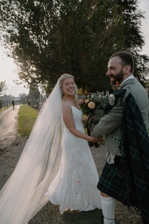 Smiling bride in white lace gown and veil holds hands with groom in Scottish tartan kilt outdoors.