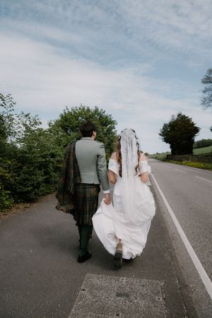 Bride and groom walking hand in hand down a country road, groom wearing a tartan kilt and bride in a white gown.