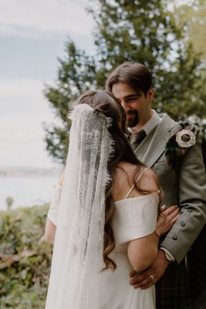 Bride and groom share a tender moment, bride in white gown with lace veil, groom in gray suit with boutonniere.