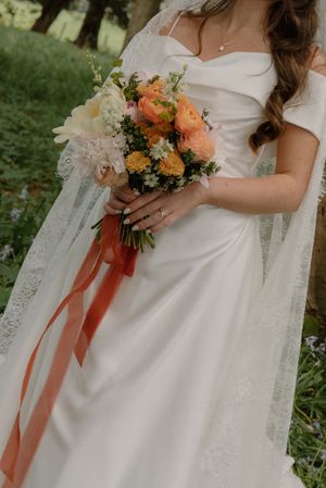 Bride in white off-shoulder gown holding colorful peach and yellow bouquet with orange ribbon outdoors.