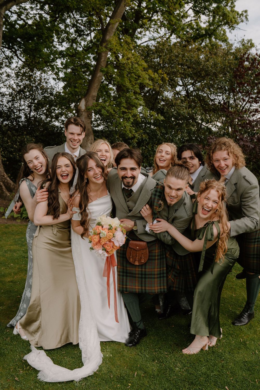 Joyful wedding party in green kilts and satin dresses celebrating outdoors, bride holding orange bouquet.