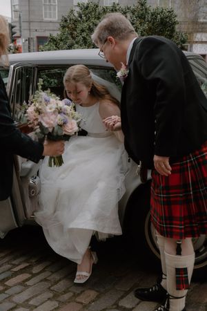 Bride in white gown exits vintage car holding bouquet, assisted by man in red tartan kilt on cobblestone street.