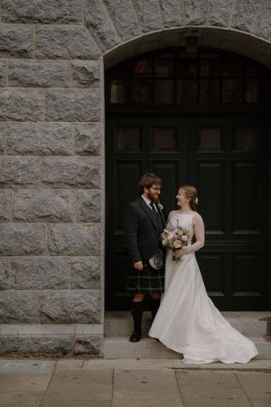 A bride and groom in Scottish kilt attire pose together in a stone archway doorway, bride holding a pastel bouquet.