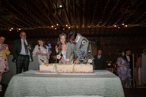 Bride and groom cutting wedding cake at rustic barn reception with guests watching in background.
