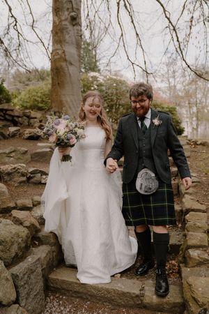 Bride in white gown and groom in green tartan kilt walk hand-in-hand down stone steps in a woodland setting.