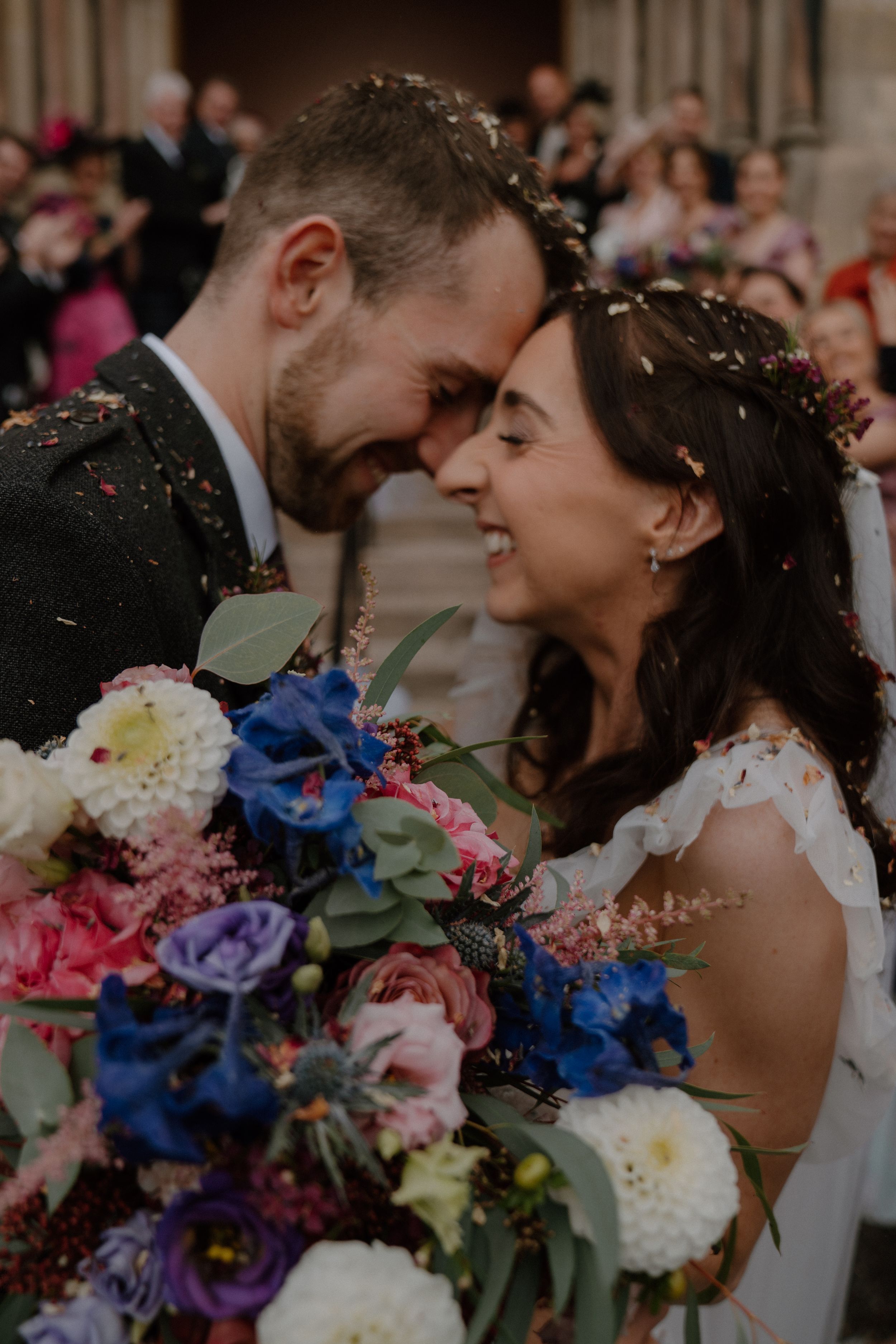 Newlywed couple touching foreheads and smiling with colorful wedding bouquet, confetti falling during outdoor ceremony.