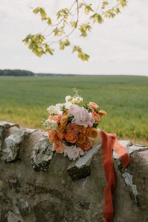 A rustic bridal bouquet of peach, orange, and white flowers with a coral ribbon rests on a stone wall overlooking green fields.