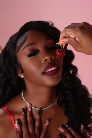 Fashion beauty portrait featuring long wavy black hair, diamond necklace, and red nail polish against pink background.