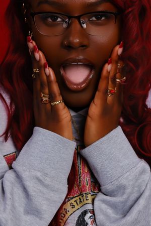 Vibrant red curly hair framing glasses, with hands adorned in multiple rings held up to cheeks in playful expression.