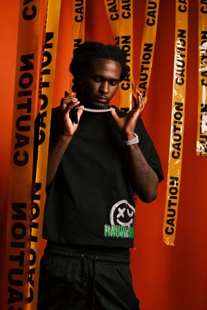 Fashion model poses against vibrant orange caution tape backdrop wearing a black t-shirt with ghost graphic design.