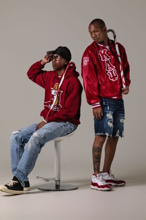 Two models wearing red varsity jackets and distressed denim on a gray backdrop for an urban fashion photoshoot.