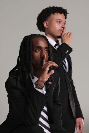 Two people in black suits and striped ties making shushing gestures against a gray background.