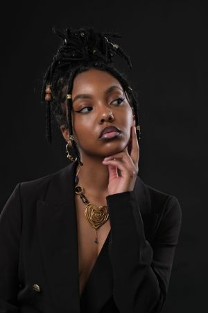 Studio portrait against dark background featuring black blazer, braided hairstyle, and gold statement necklace.