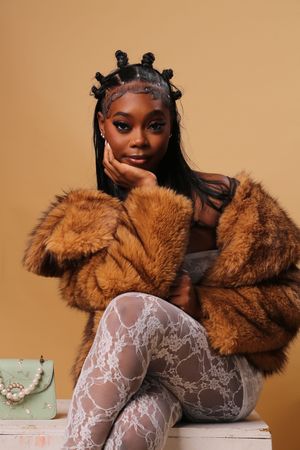 Fashion portrait with brown fur coat, delicate lace tights, and stylish bantu knots hairstyle against beige backdrop.