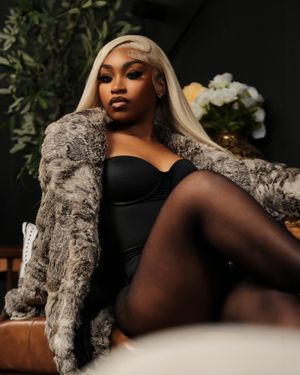 Glamorous portrait in a plush grey fur coat and black bodysuit against a dark background with greenery and white flowers.