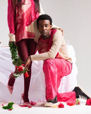 Person in red and cream colored clothing poses against white backdrop with scattered red flowers and greenery.