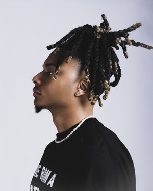 Profile view shot in studio lighting showing stylish dreadlocks hairstyle and black top with pearl necklace against gray background.