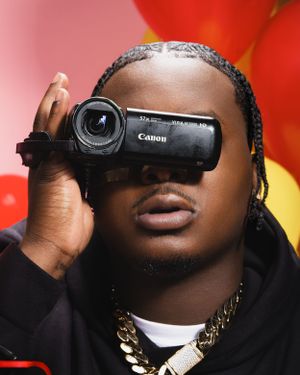 A person holds a Canon video camera up to their face while wearing a black hoodie and gold chain against a red background.
