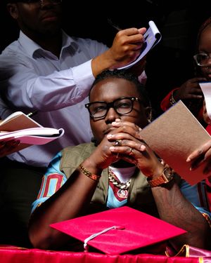 A graduate student in glasses wears a green jacket and thoughtfully rests their chin on clasped hands near a pink graduation cap.