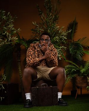Person relaxing on a wicker chair surrounded by tropical plants in a moody studio setting with warm lighting.