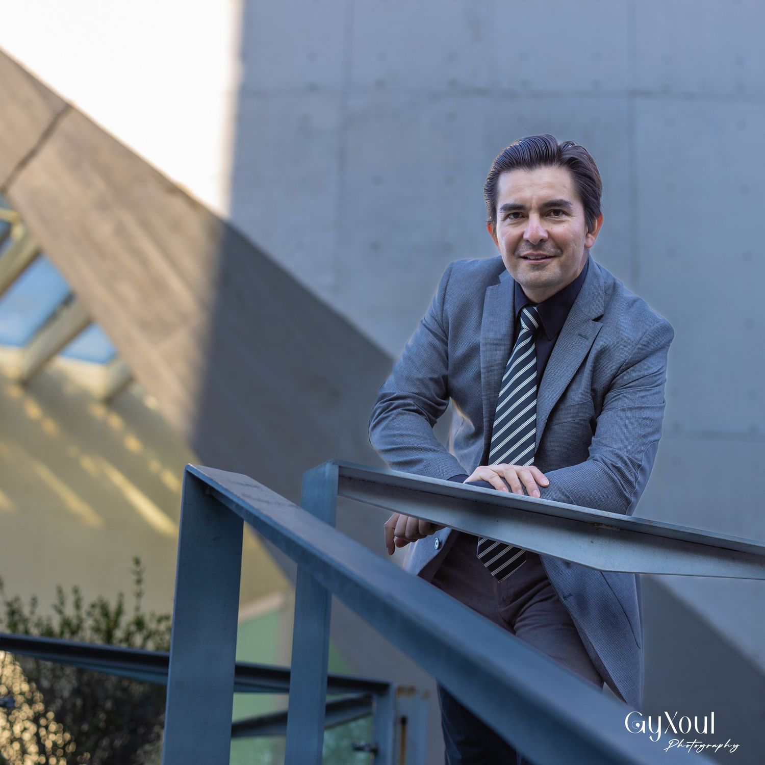Professional in gray suit and tie poses on outdoor stairway railing.