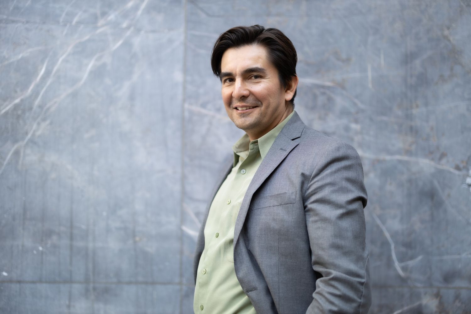 Man in business attire smiles warmly against textured gray wall background.