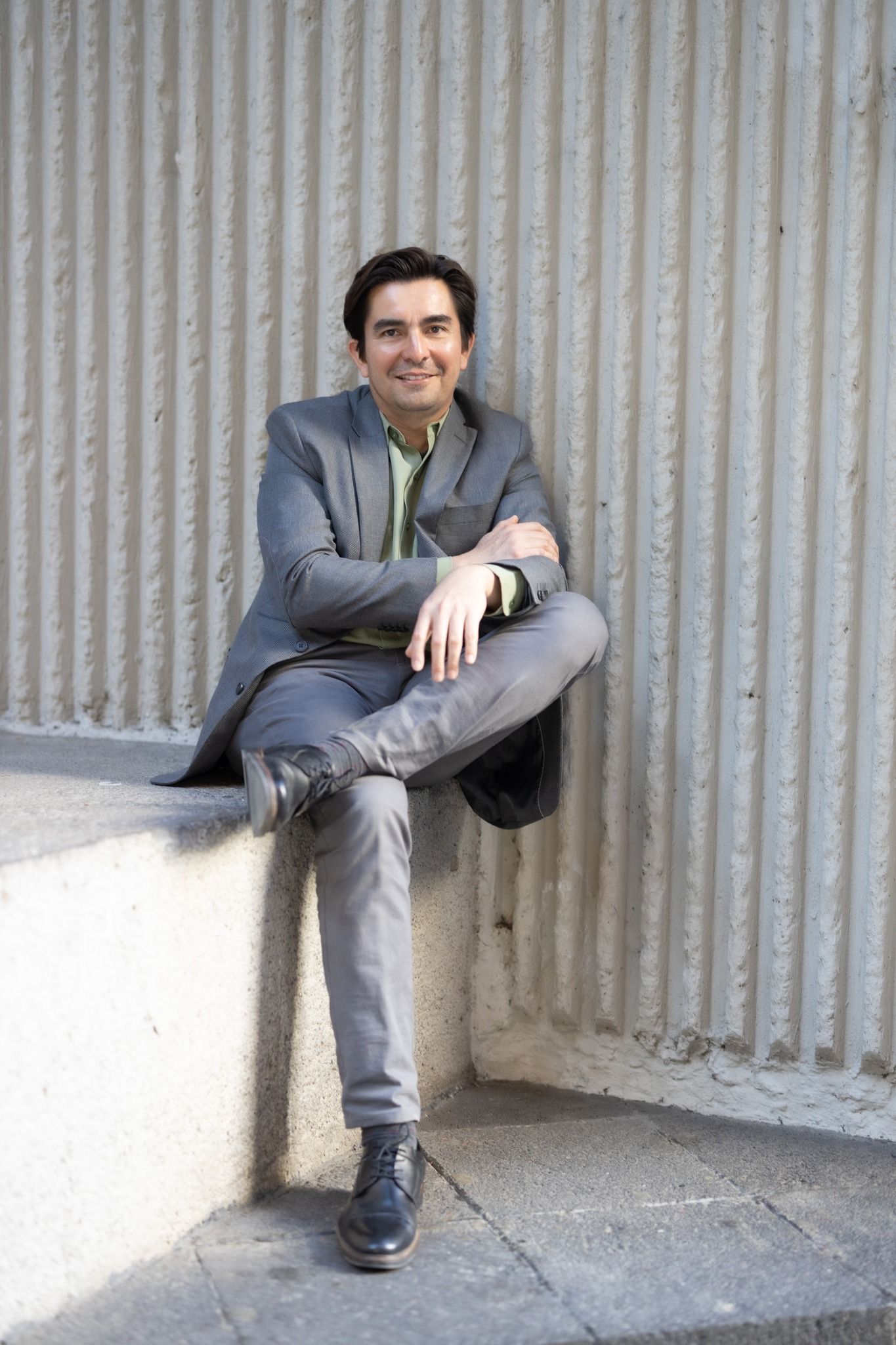Professional in gray suit sits casually on concrete ledge against ribbed wall.