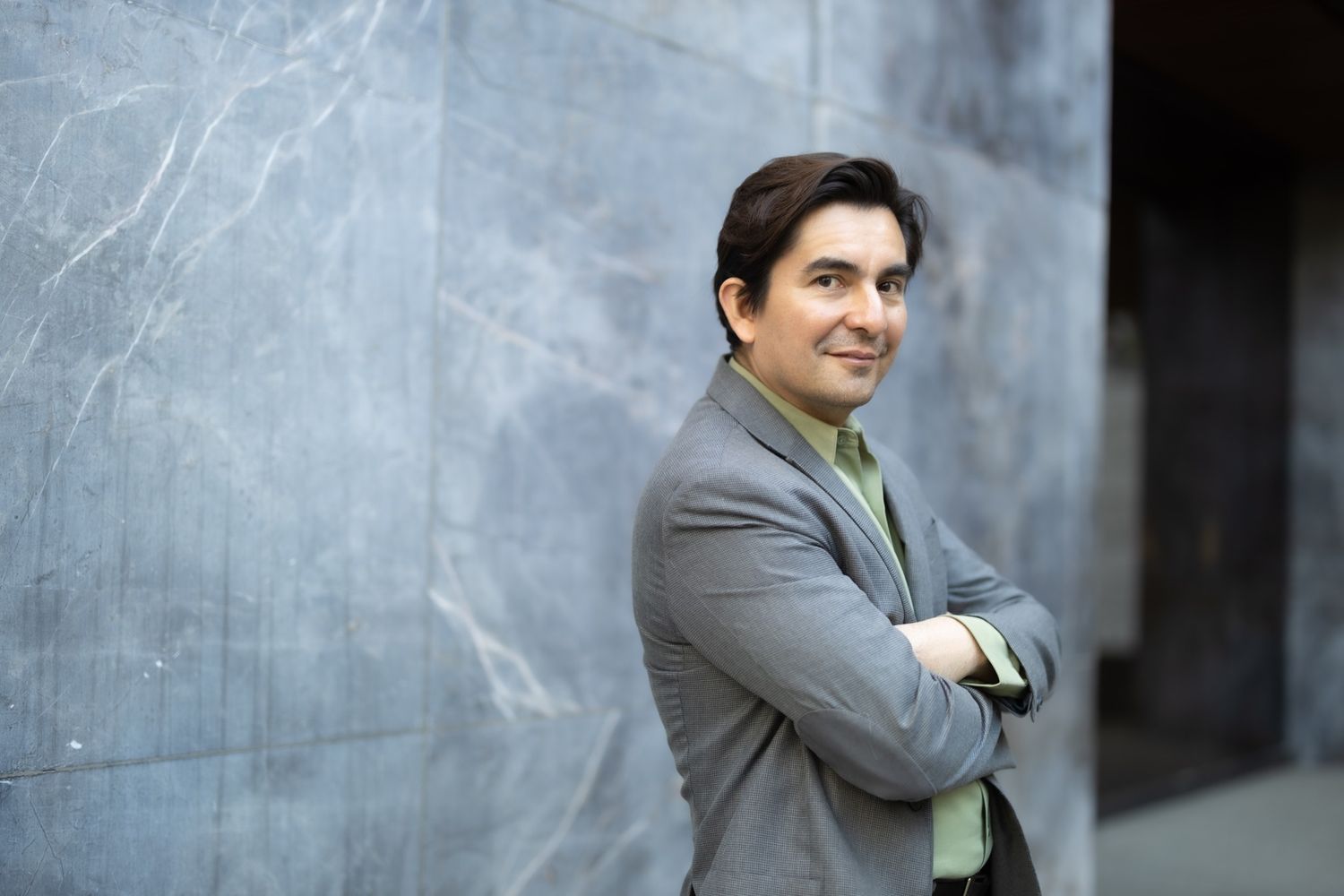 Businessman in gray suit poses confidently against concrete wall.