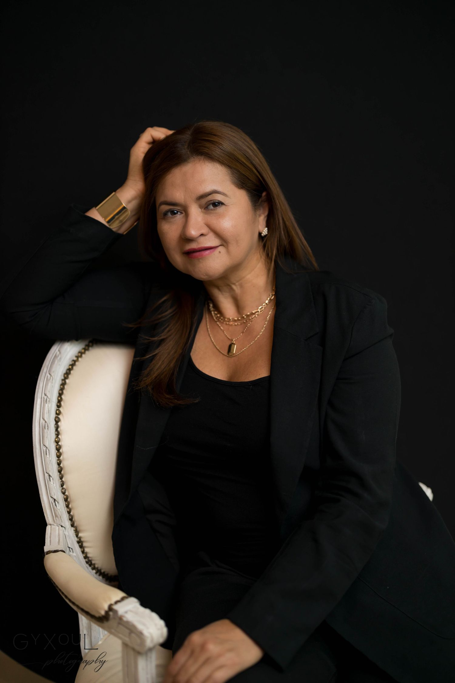 Studio portrait of someone in black attire seated on light colored chair.