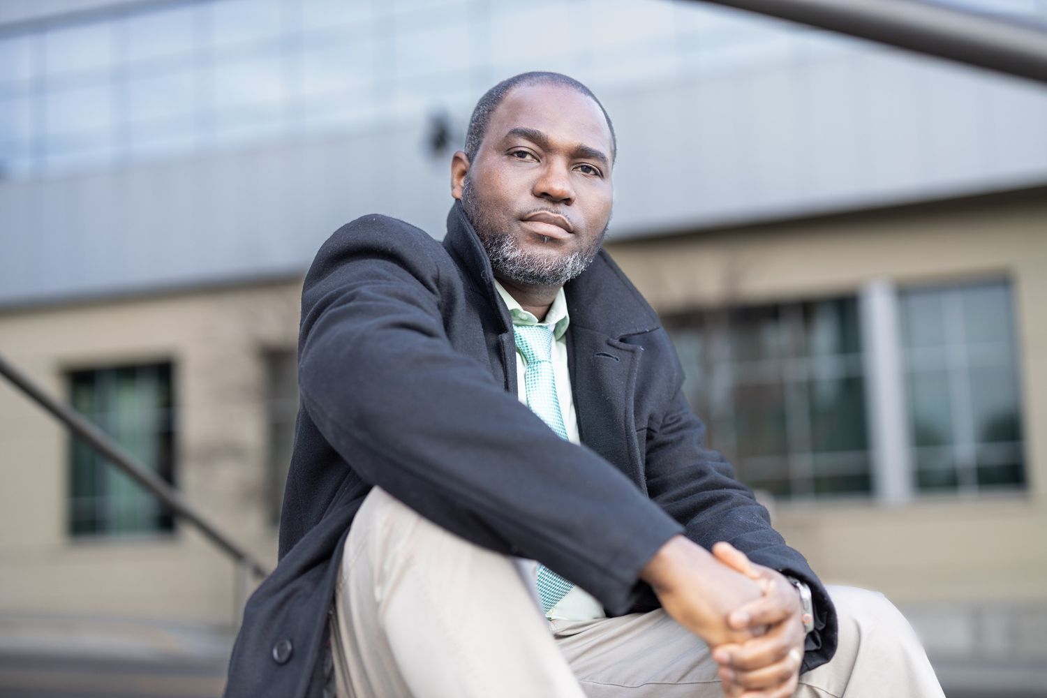 Outdoor portrait showing someone in a dark jacket seated against an architectural backdrop.