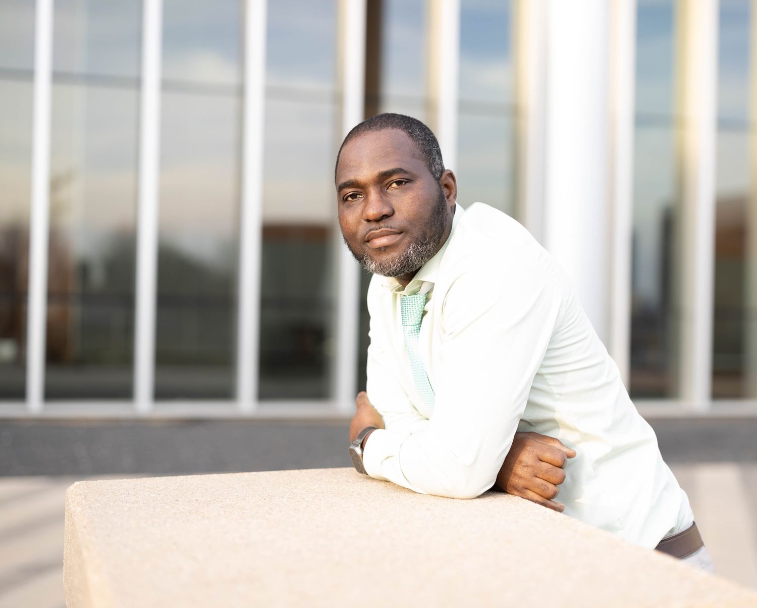 Man in white blazer leans on outdoor ledge with modern building in background.