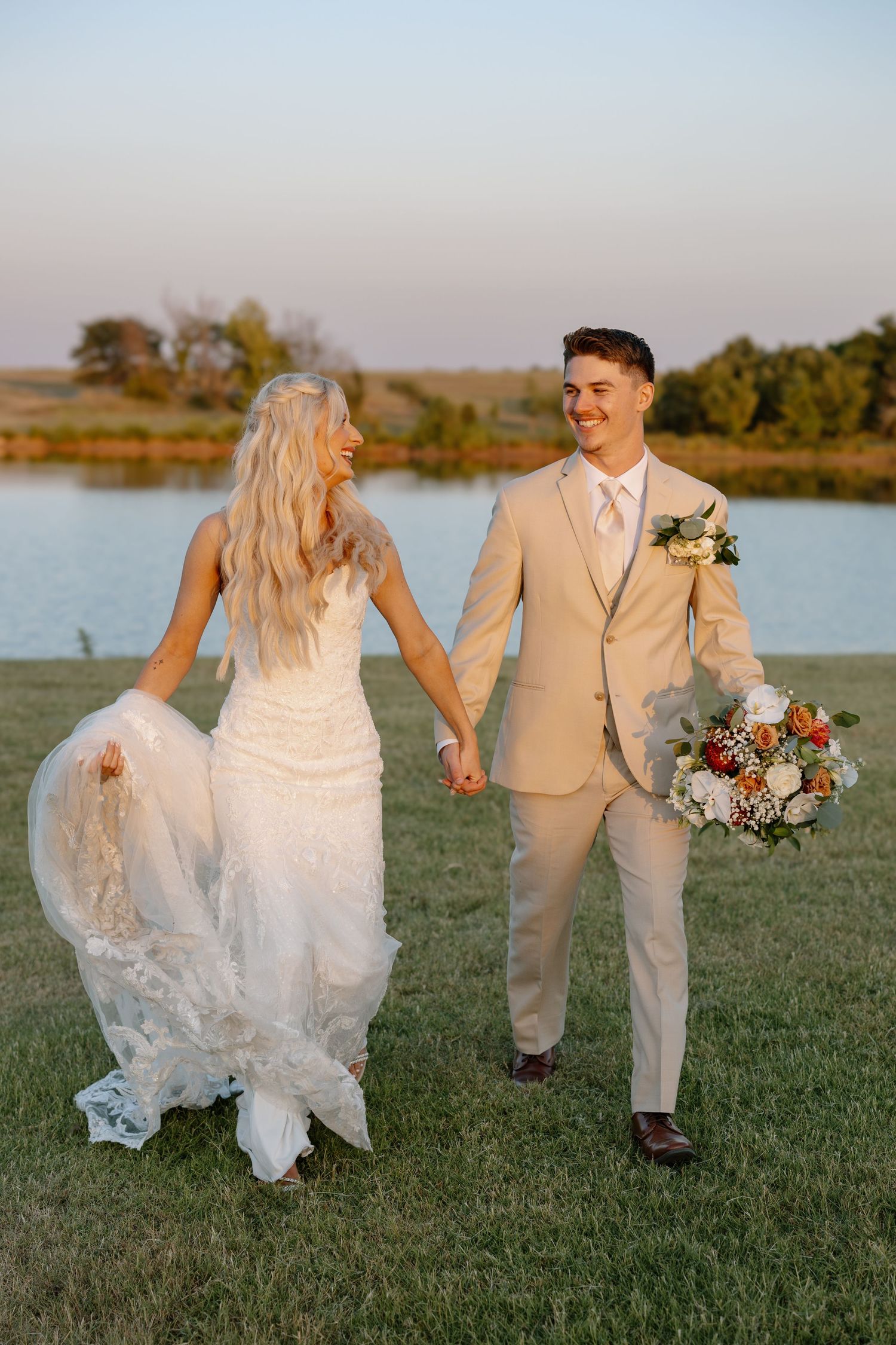 Newlywed couple laughing while walking on grass by lake, bride in white dress and groom in tan suit.